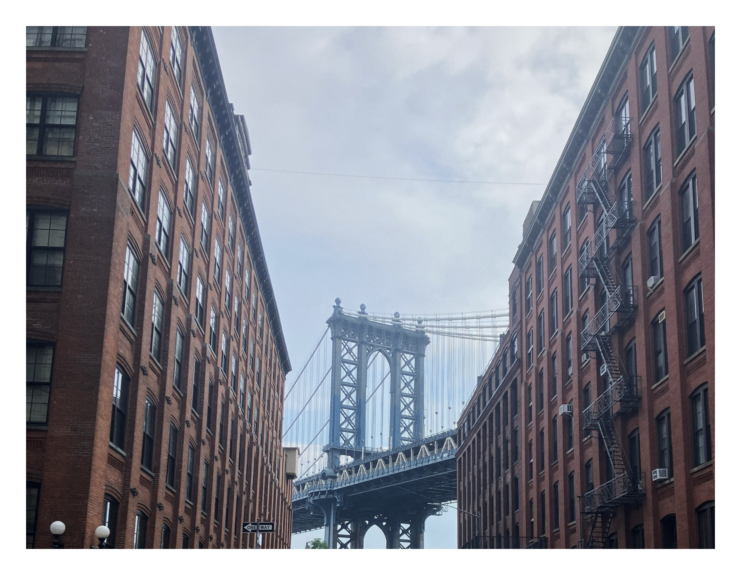photo of the Manhattan Bridge, constructed of blue-grey steel latticework, framed on either side by tall red-brick warehouse buildings
