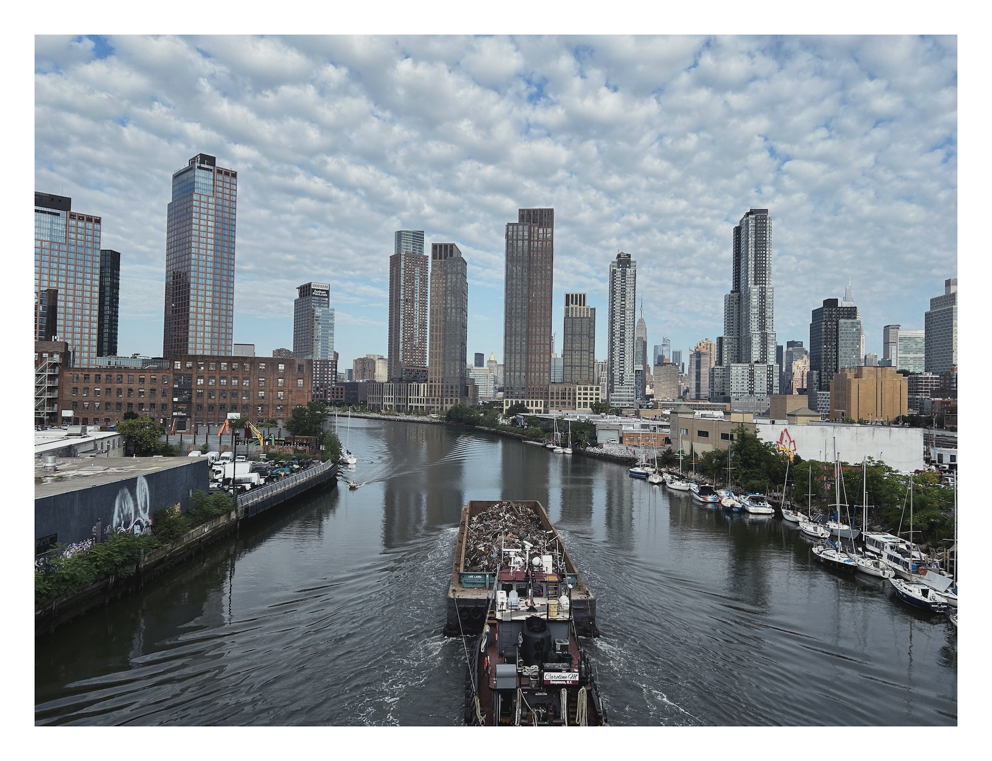 a barge carrying a load of rusty scrap metal travels down a creek lined with warehouses toward a city skyline in the distance
