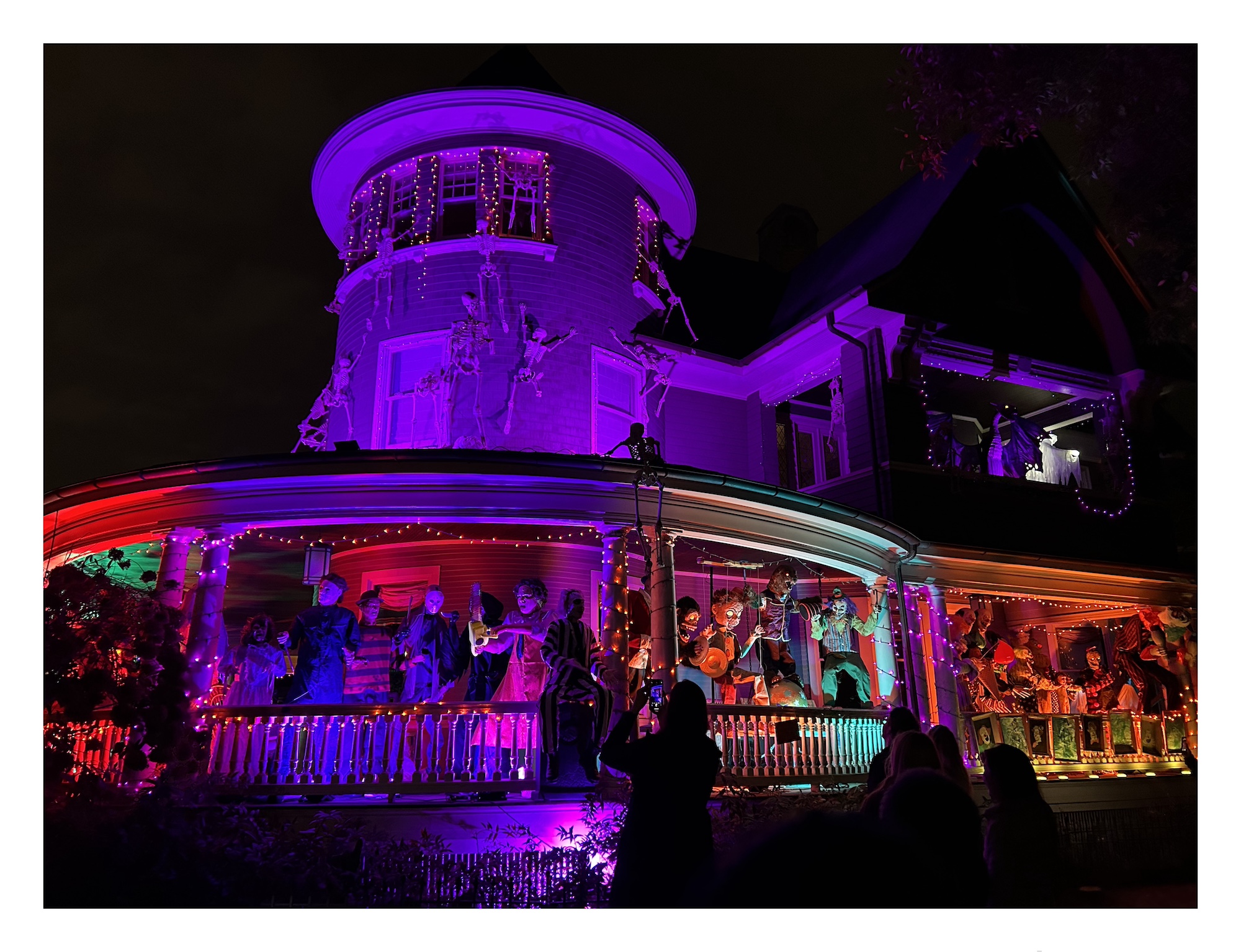 nighttime scene of a large Victorian home covered with Halloween decorations –&nbsp;skeletons, statues, animatronic monsters&nbsp;– and glowing with dramatic purple and orange lighting