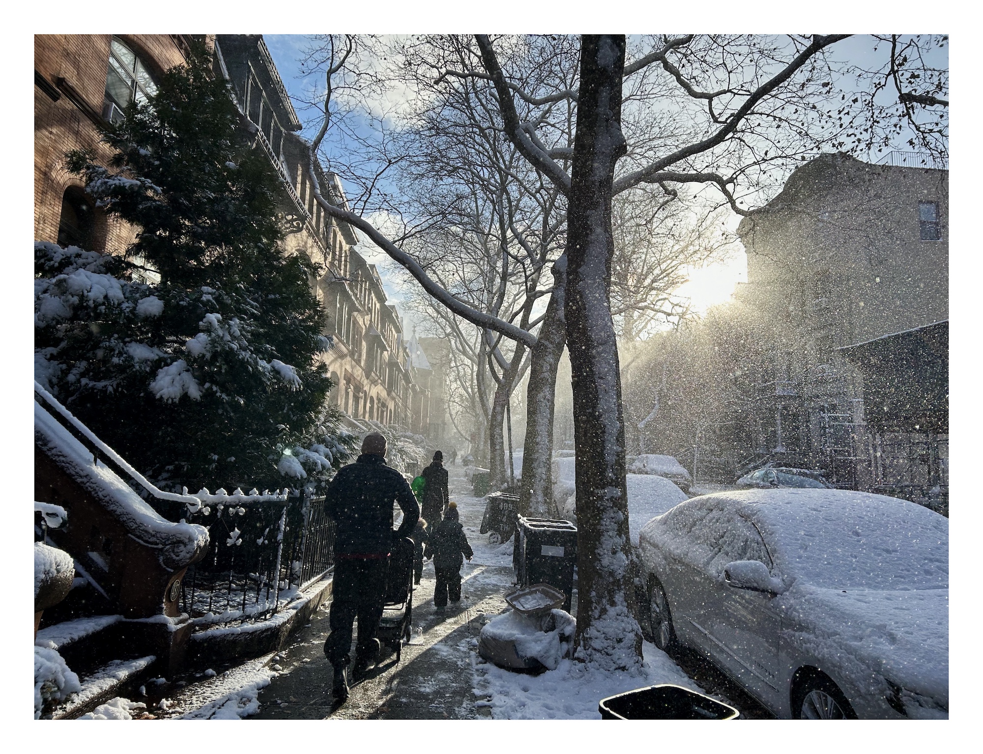 a man pushes a stroller up a narrow snow-covered sidewalk, as two children and a woman walk in front. the sun is low in the sky and tiny snow flurries scatter the early morning light