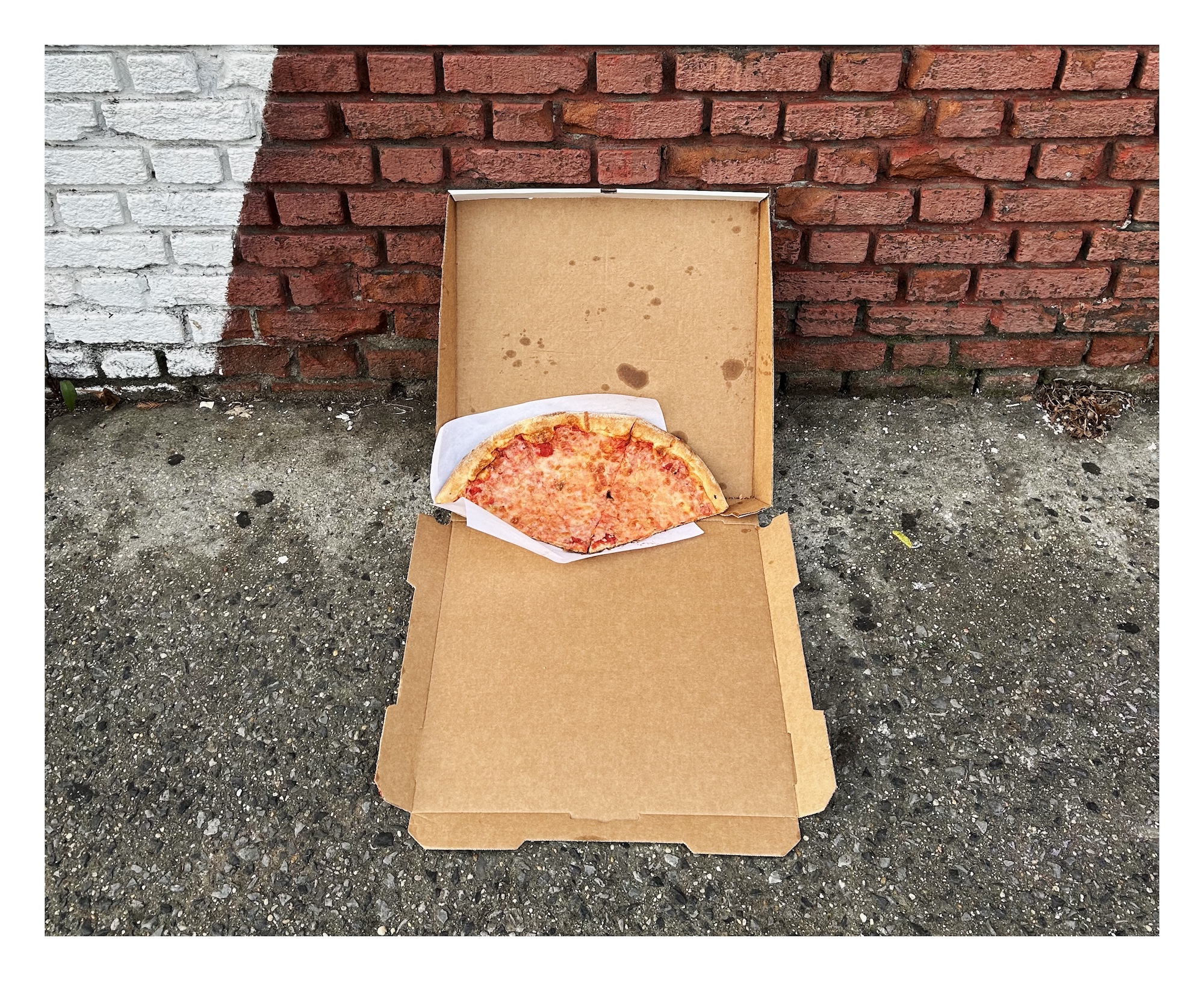 An open pizza box – with three cheese slices – lies on a sidewalk, with its lid propped against a brick wall.