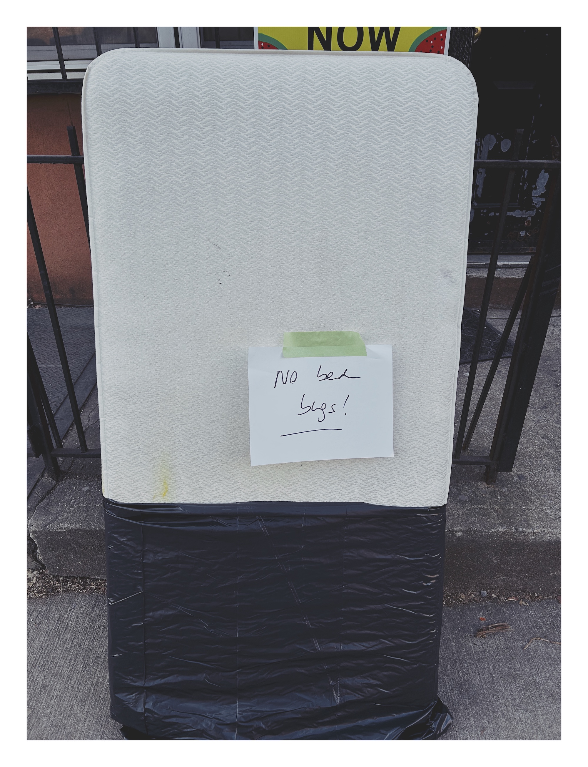 A small mattress, half-wrapped in black plastic, propped again a fence. There is a paper sign taped to the mattress that says 'No bed bugs!'