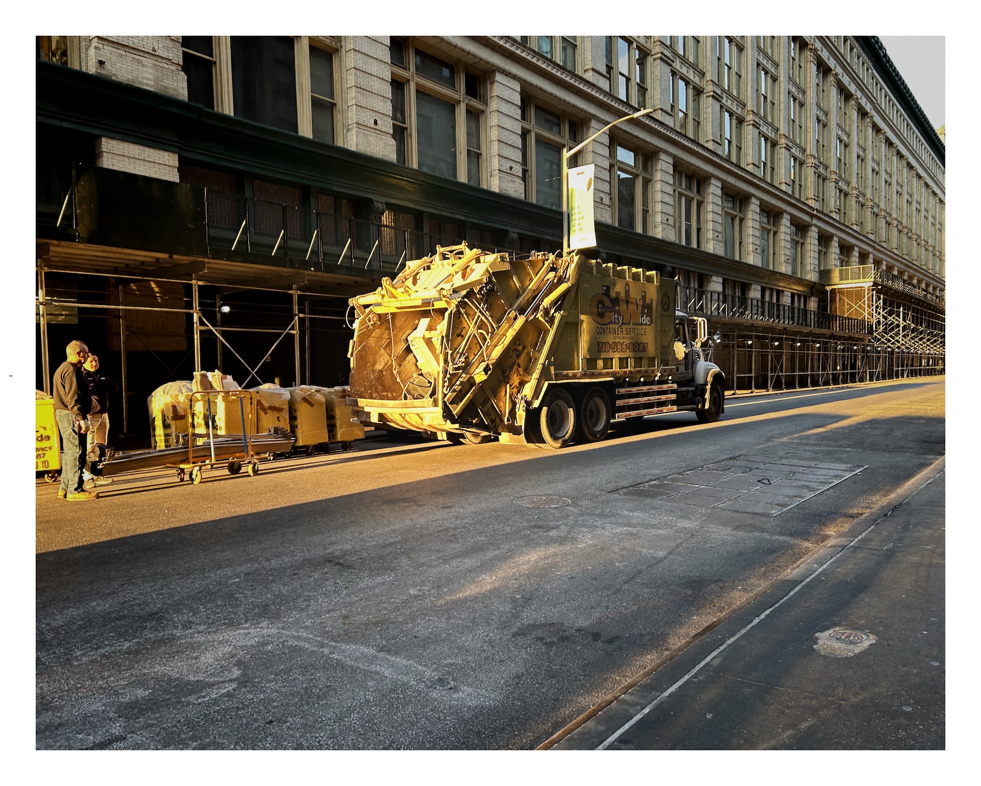 a narrow band of warm yellow morning sunlight filters down a Manhattan street and falls on two men standing near a yellow garbage truck
