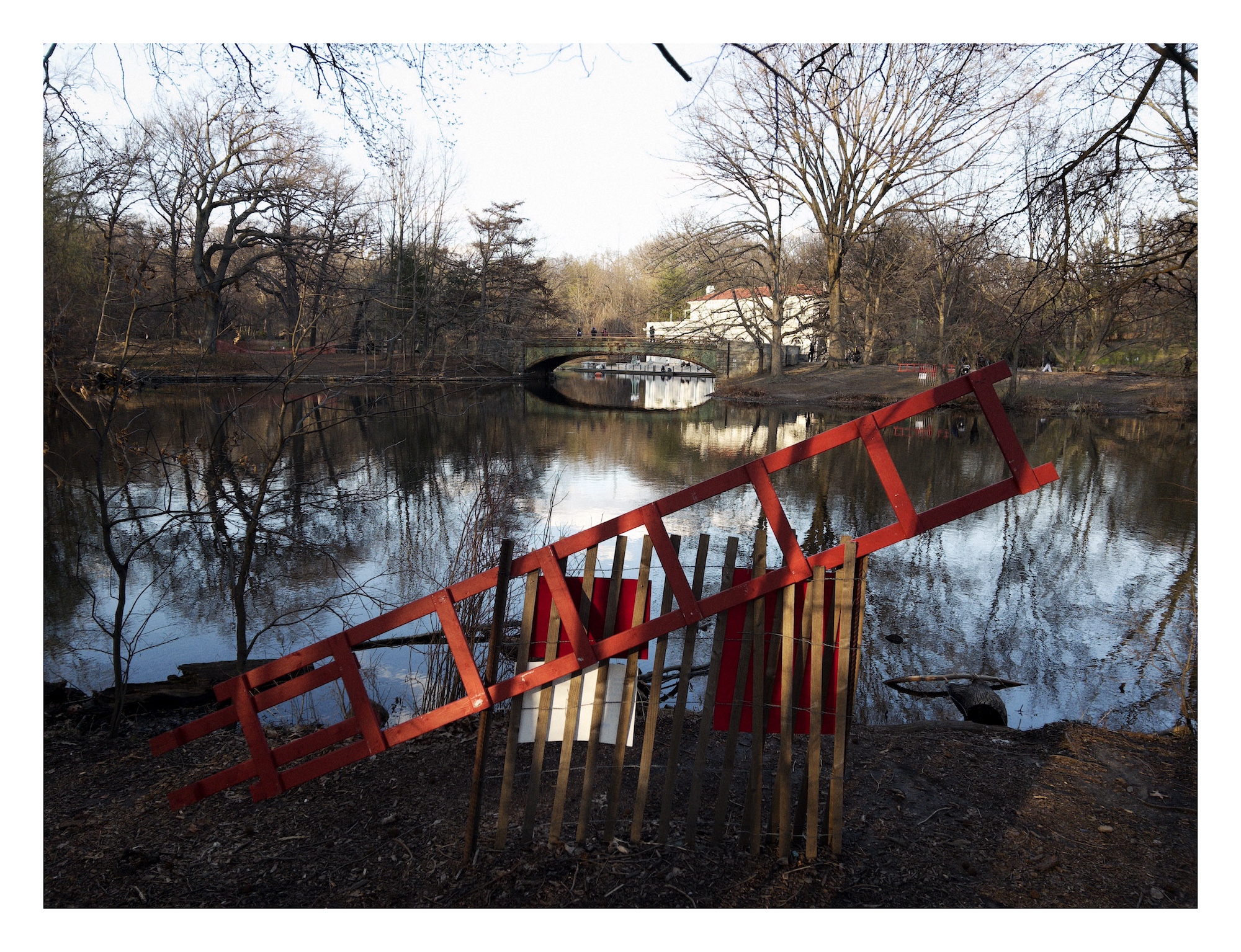 a red rescue ladder rests propped against a wooden fence alongside a pond; a bridge and boathouse are on the far side of the pond