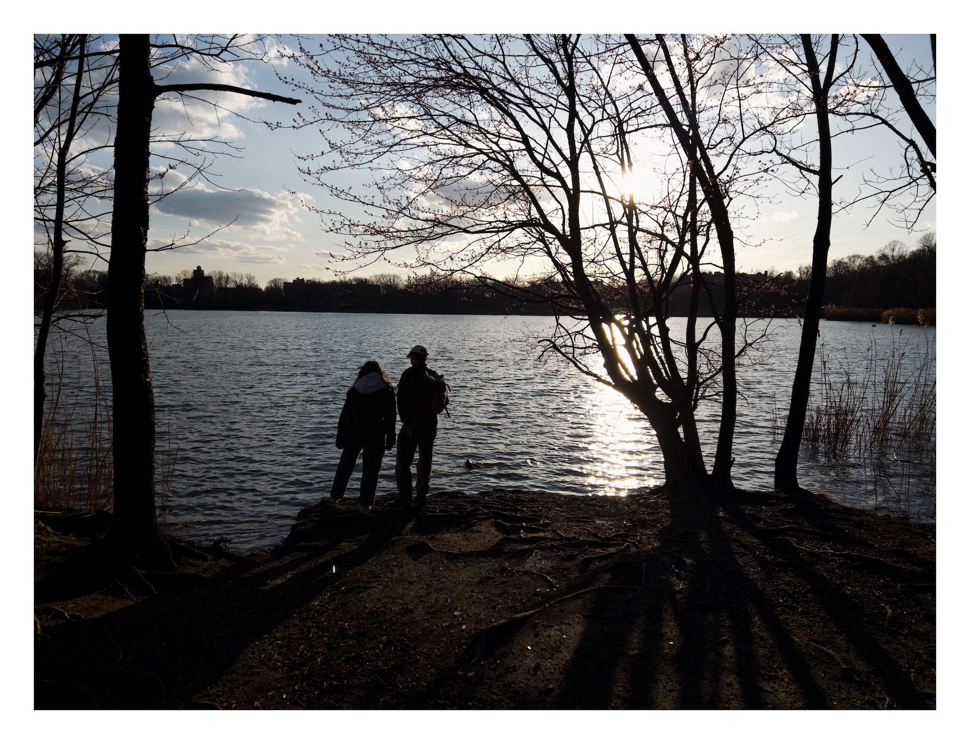 a woman and man scene from behind as they stand by the shore of a lake; the are silhouetted by the sun setting behind the far shore