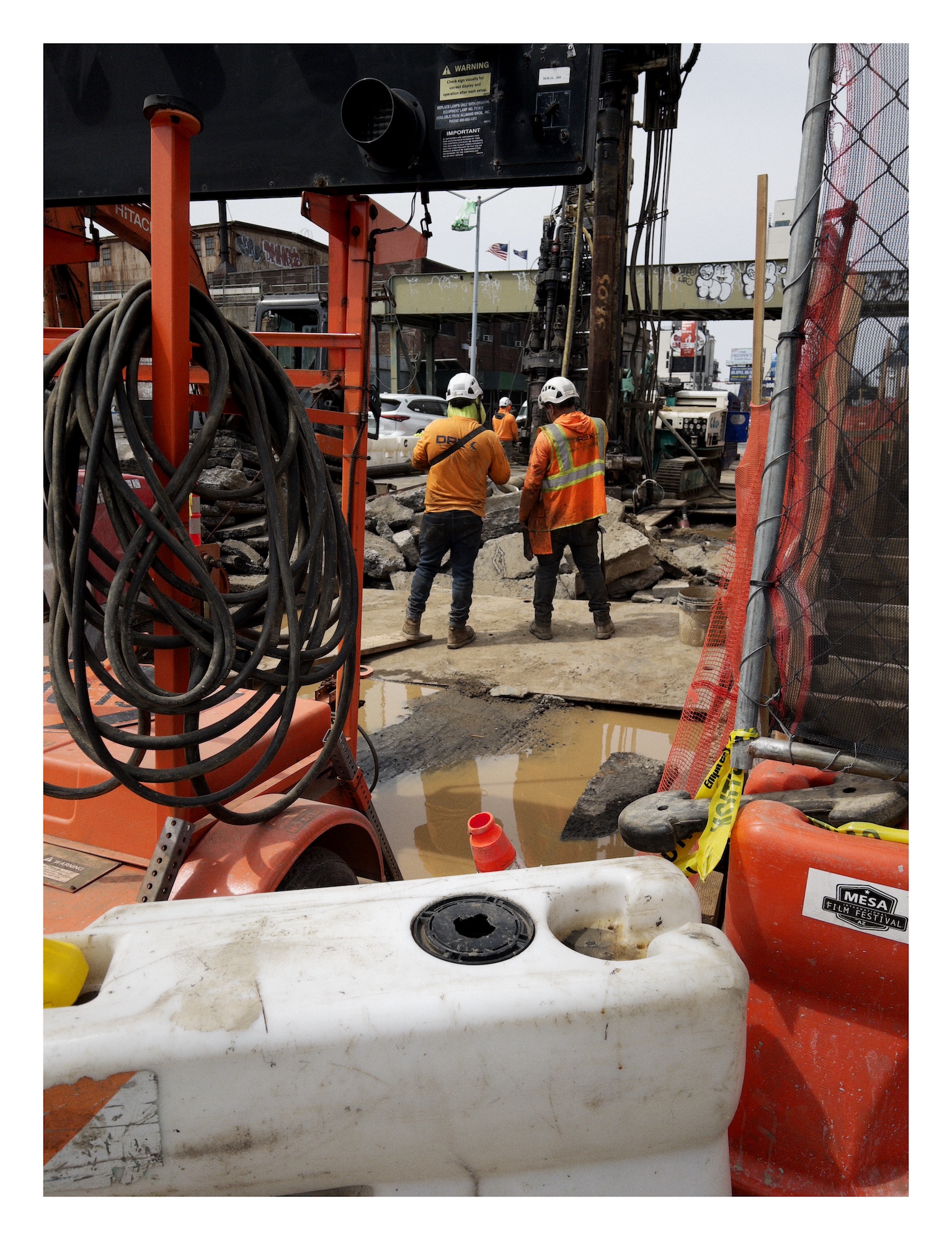 men wearing orange shirts and constructions vests work on a road repair project surrounded by orange cones and orange barriers and a sign posted on an orange frame