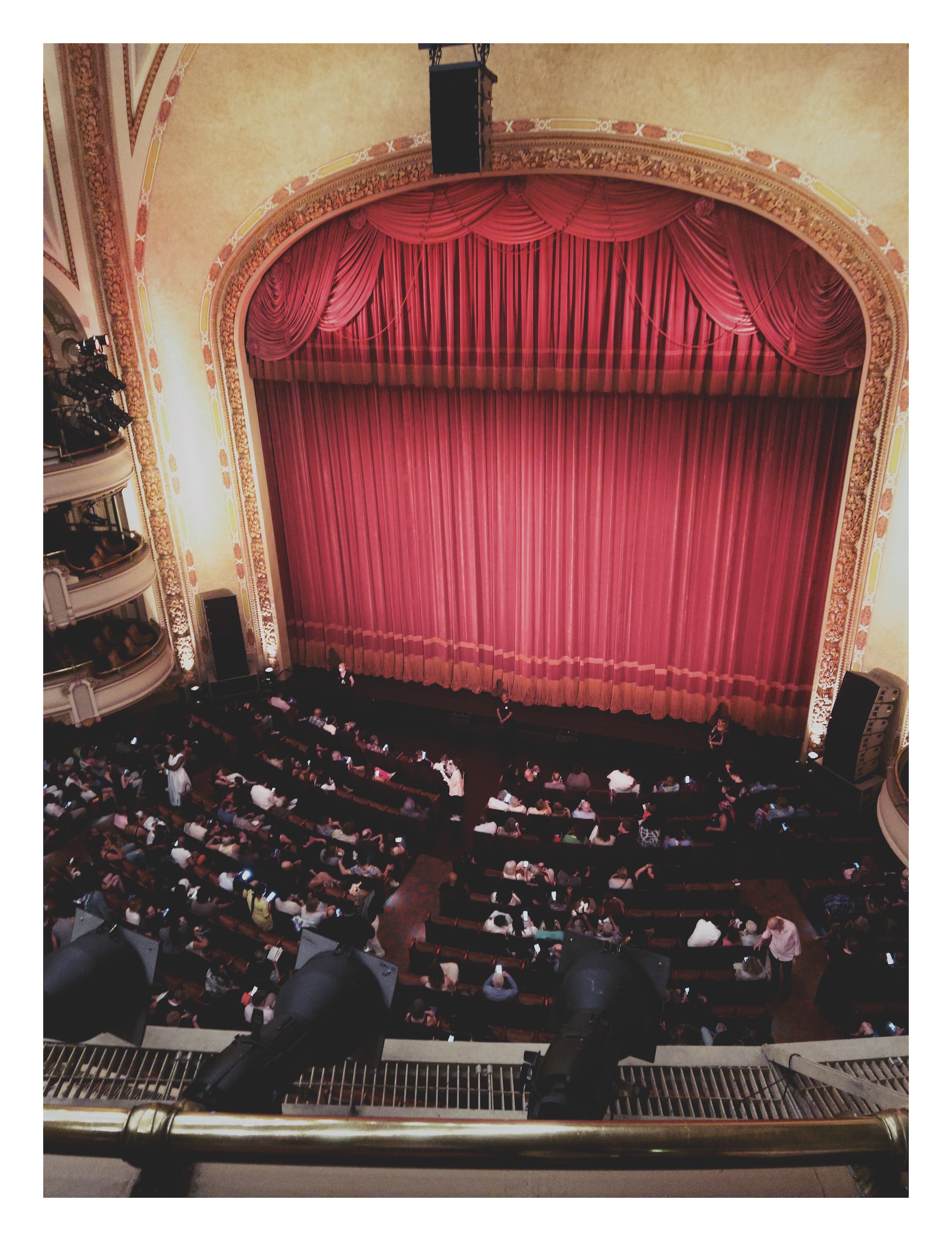 a view from the balcony of an ornate opera house, looking down toward the audience taking their seats in front of the main stage, closed off by tall red curtains