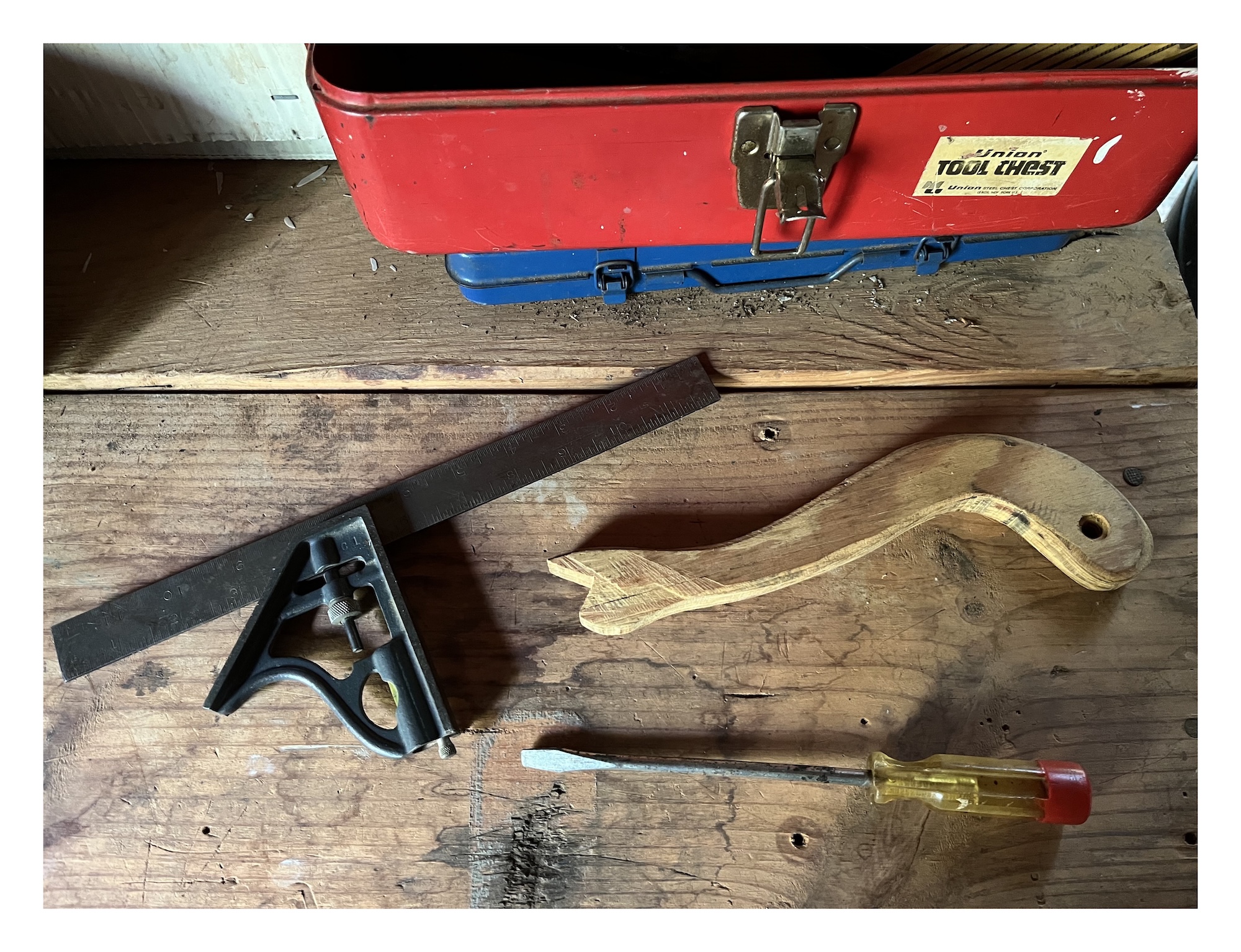 three tools on a workshop table: a metal combo square, a notched wooden handle guide, and a screwdriver
