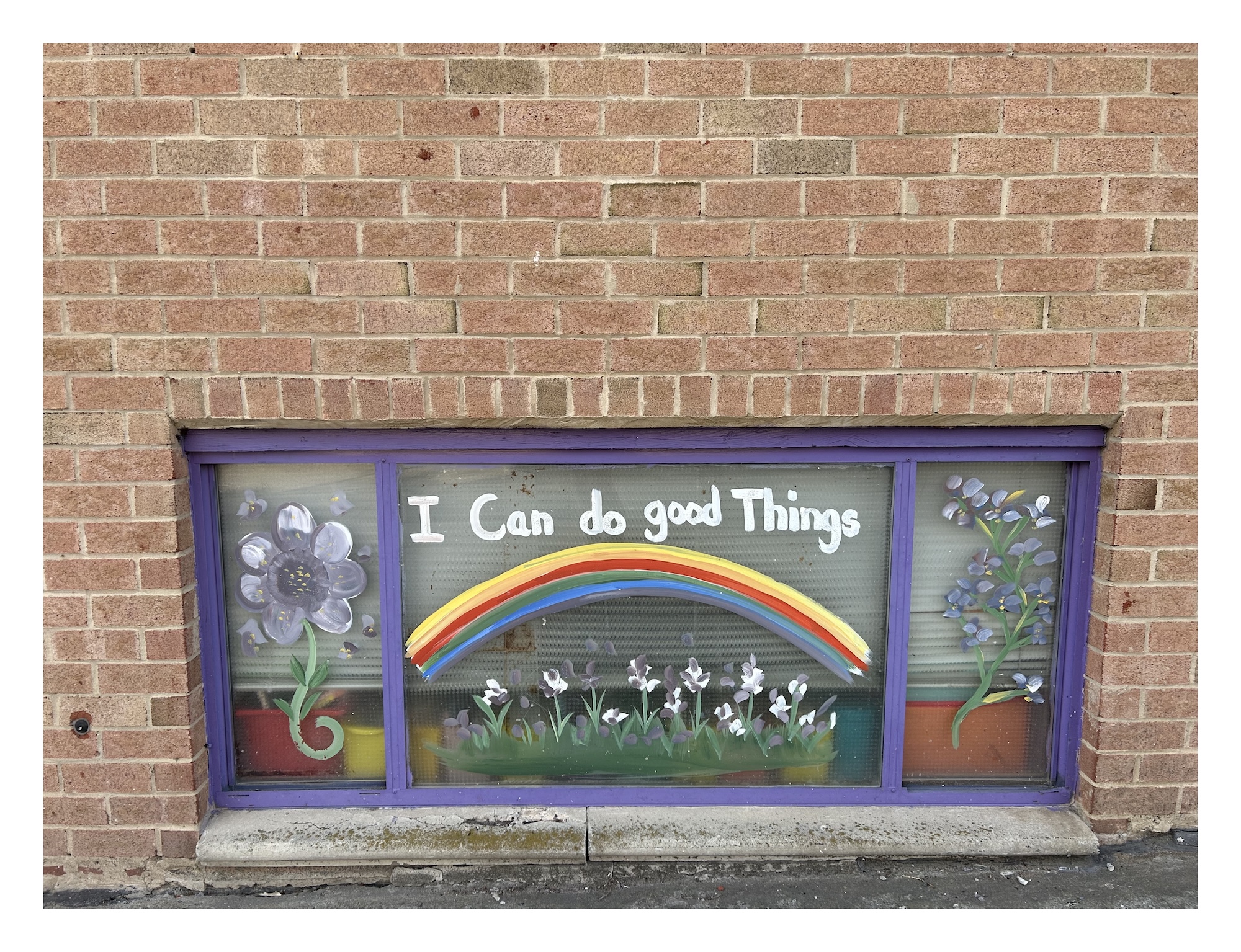 a ground-level window in a brick school building painted with violet flowers, a rainbow, and the words "I can do good things"