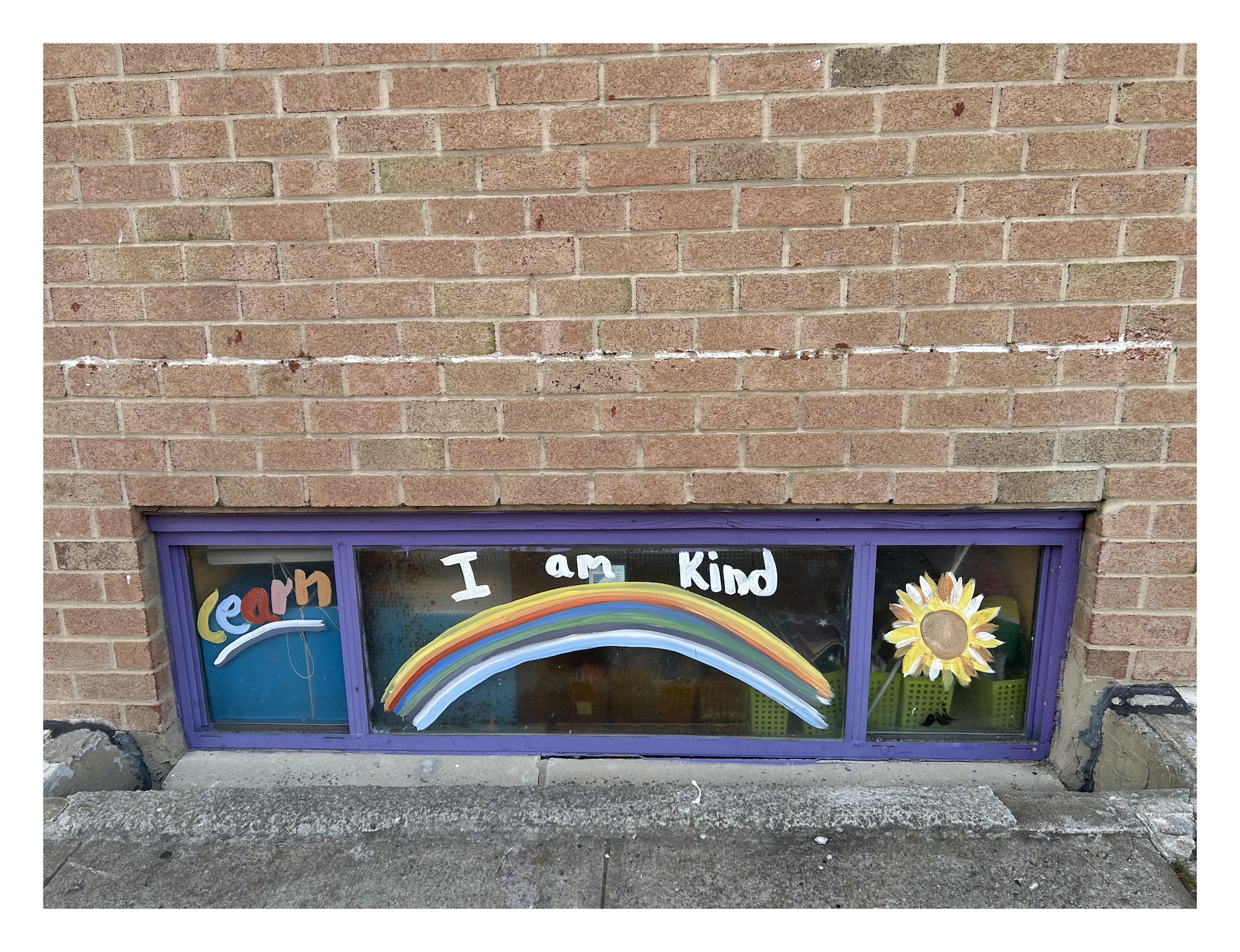 a ground-level window in a brick school building painted with a sunflower, a rainbow, and the words "I am kind"