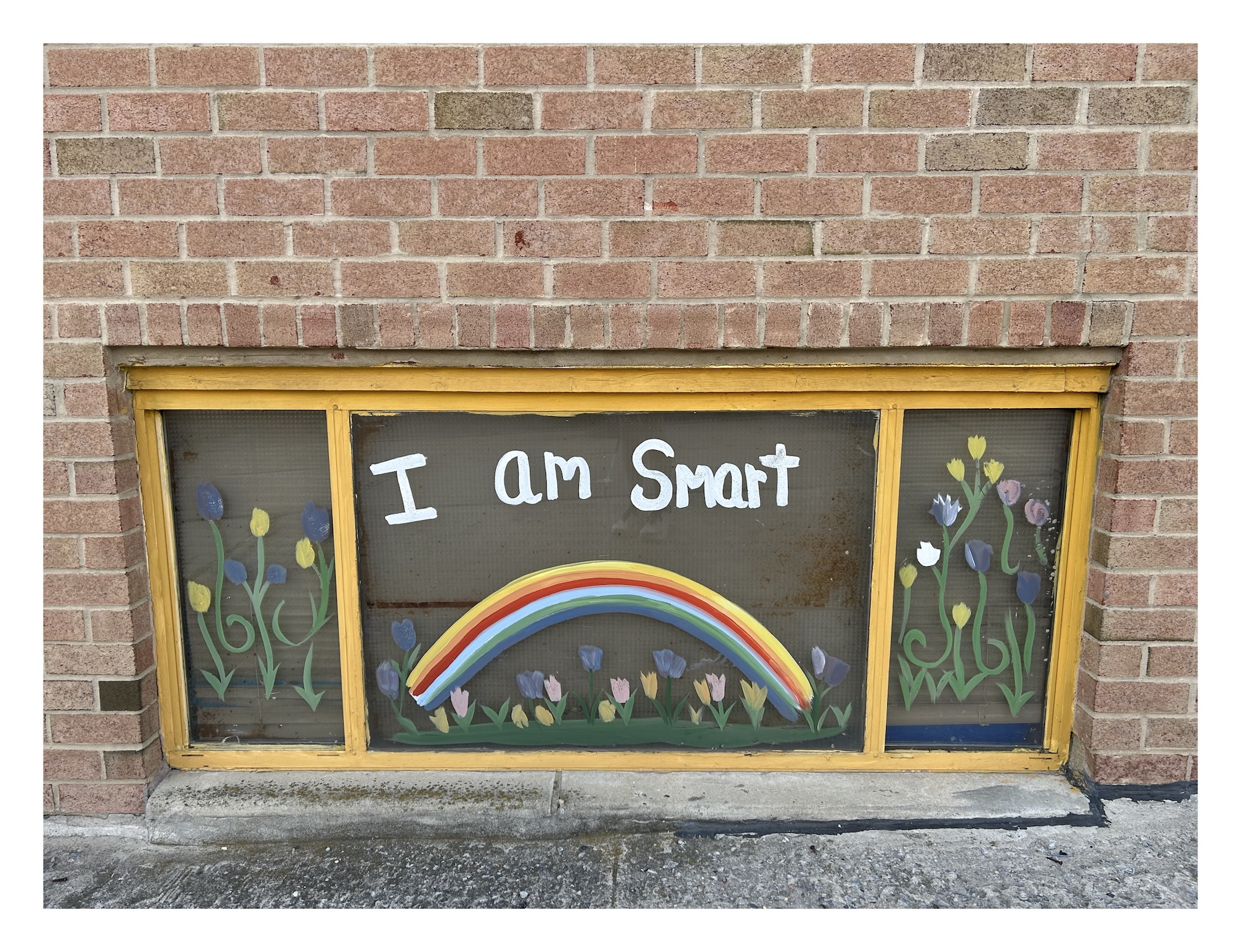 a ground-level window in a brick school building painted with flowers, a rainbow, and the words "I am smart"