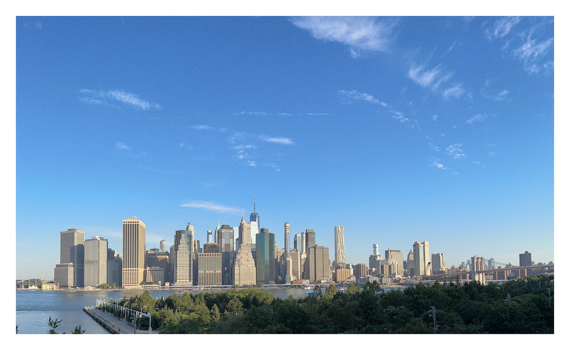 next to a river, tall skyscrapers buildings pierce a big blue sky; view of lower Manhattan skyline from Brooklyn Promenade