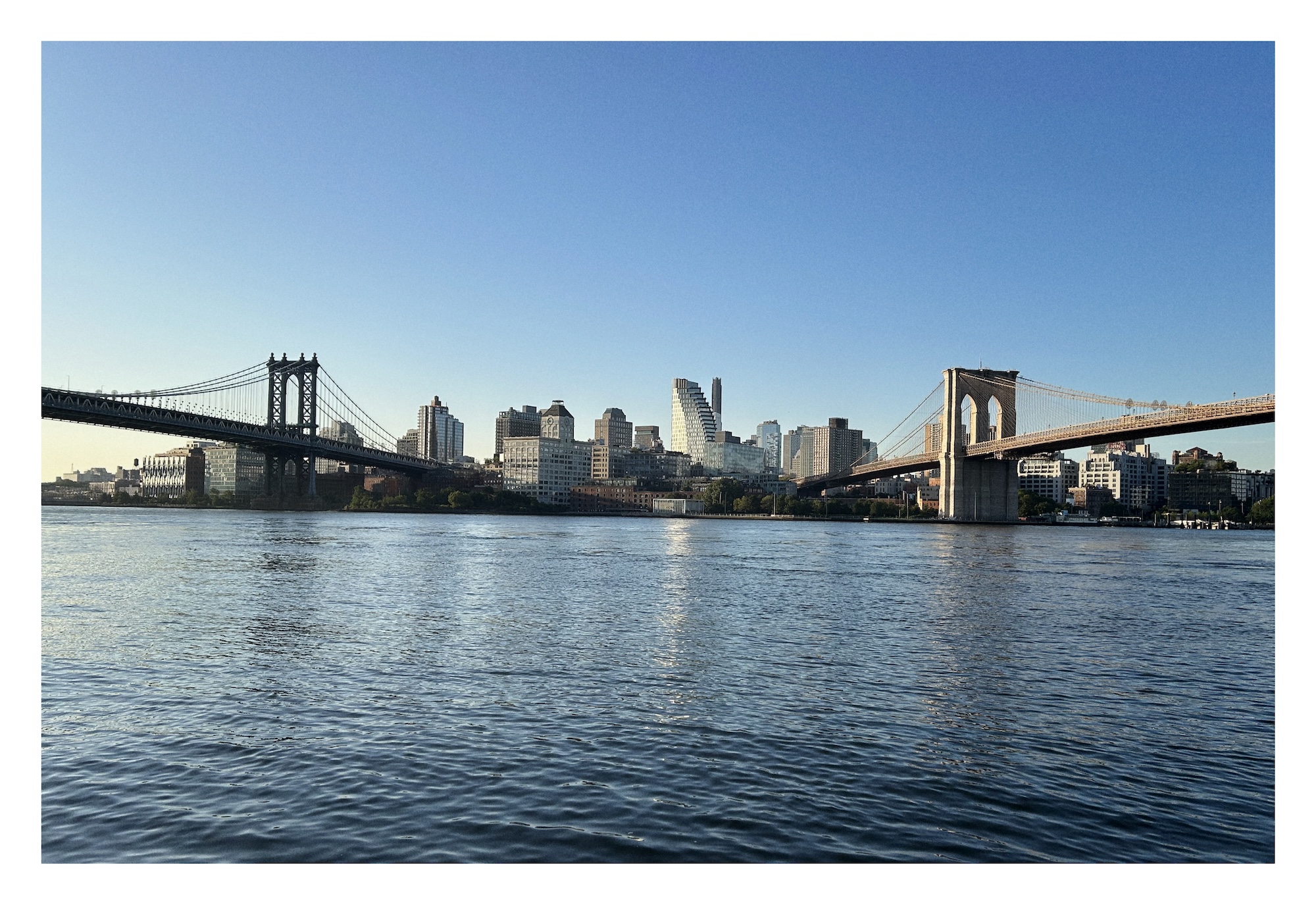 view from Manhattan over the dark blue water of the East River looking at the dark blue steel Manhattan Bridge on the left and the granite beige Brooklyn Bridge on the right, with downtown Brooklyn skyscraper in between
