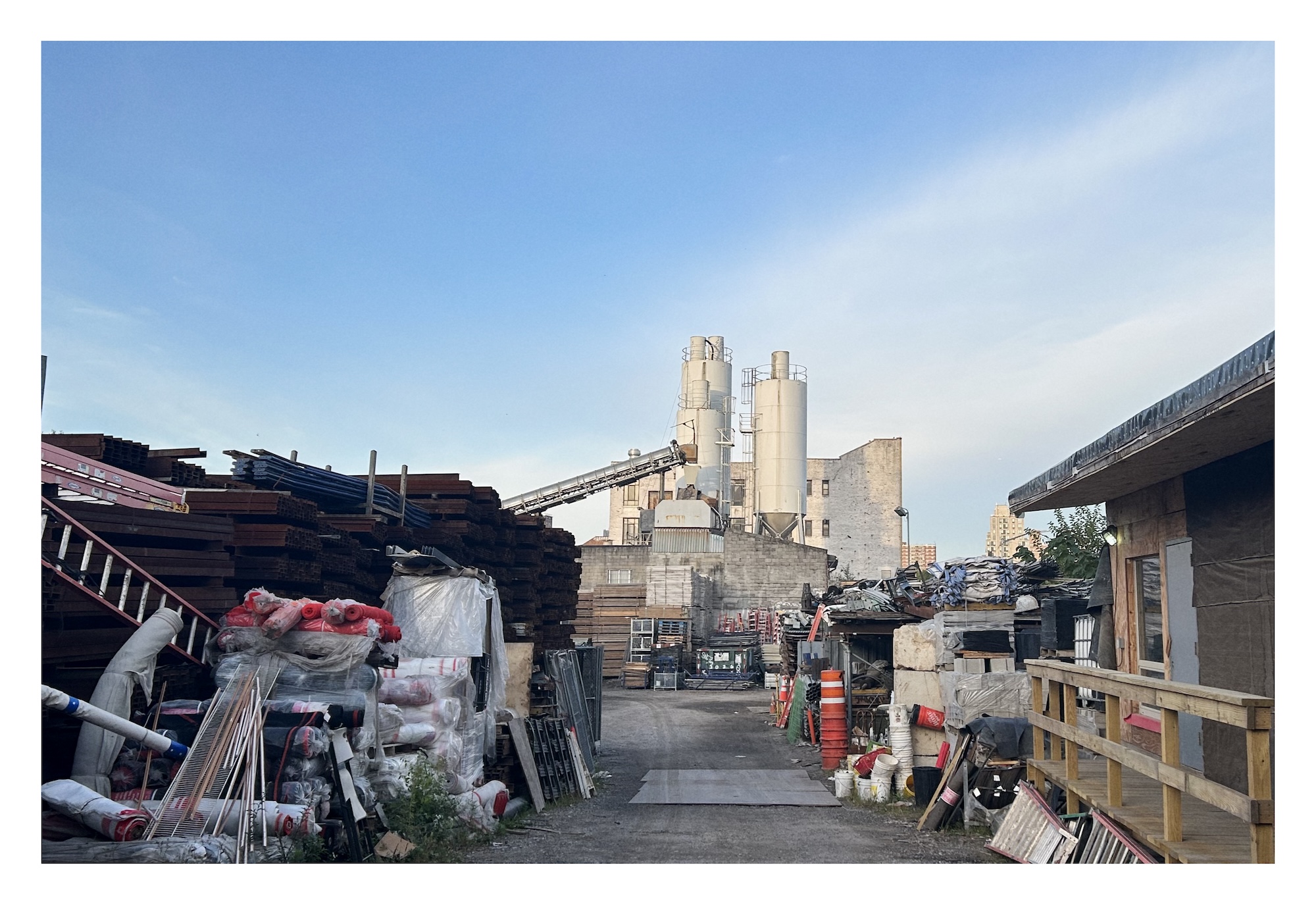 view down a hardpark dirt road between stacks of lumber and industrial equipment, with tall beige factory buildings and silos loom against a blue sky