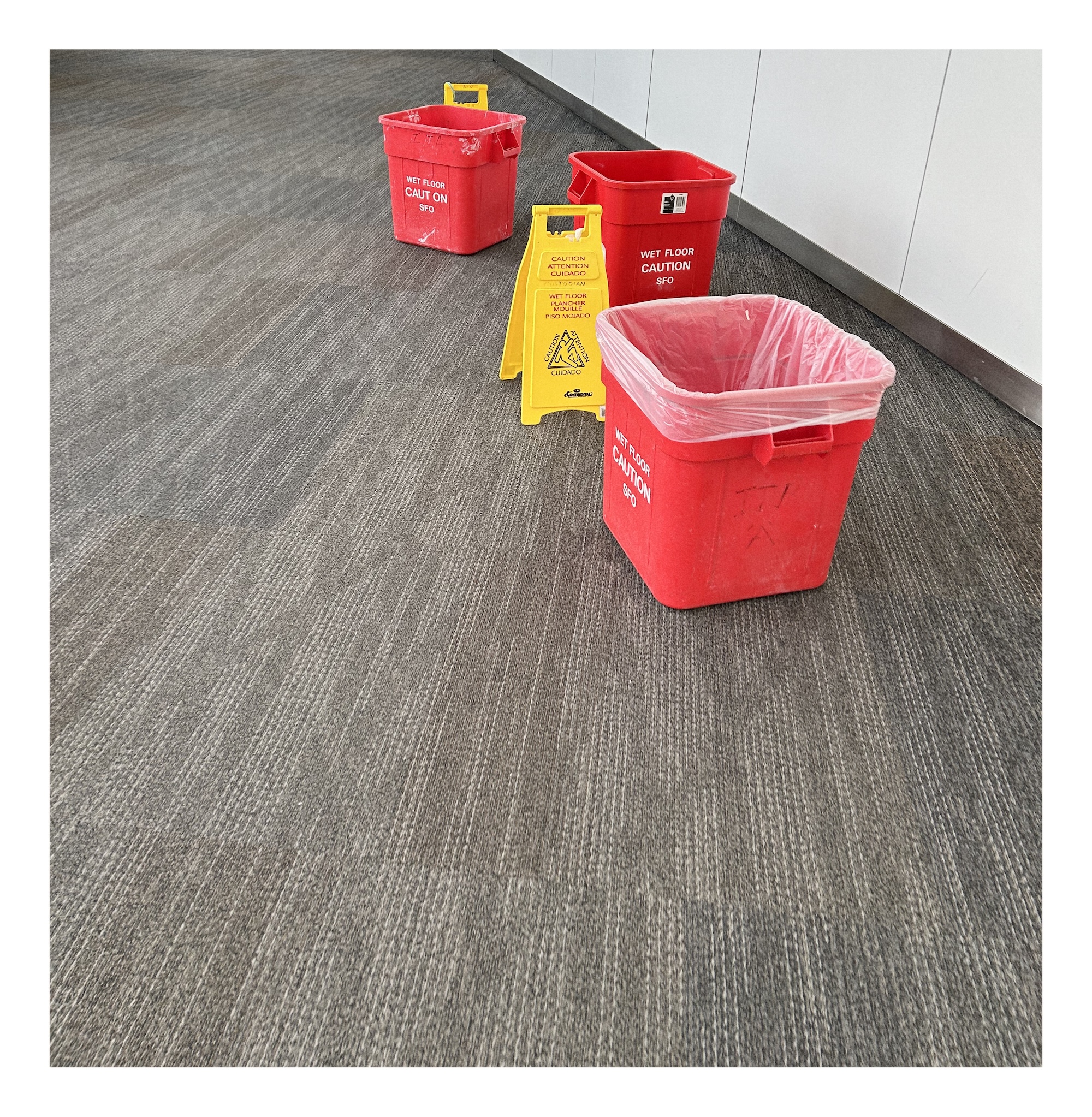 three large square red buckets and two yellow caution signs place on a bland airport grey carpet
