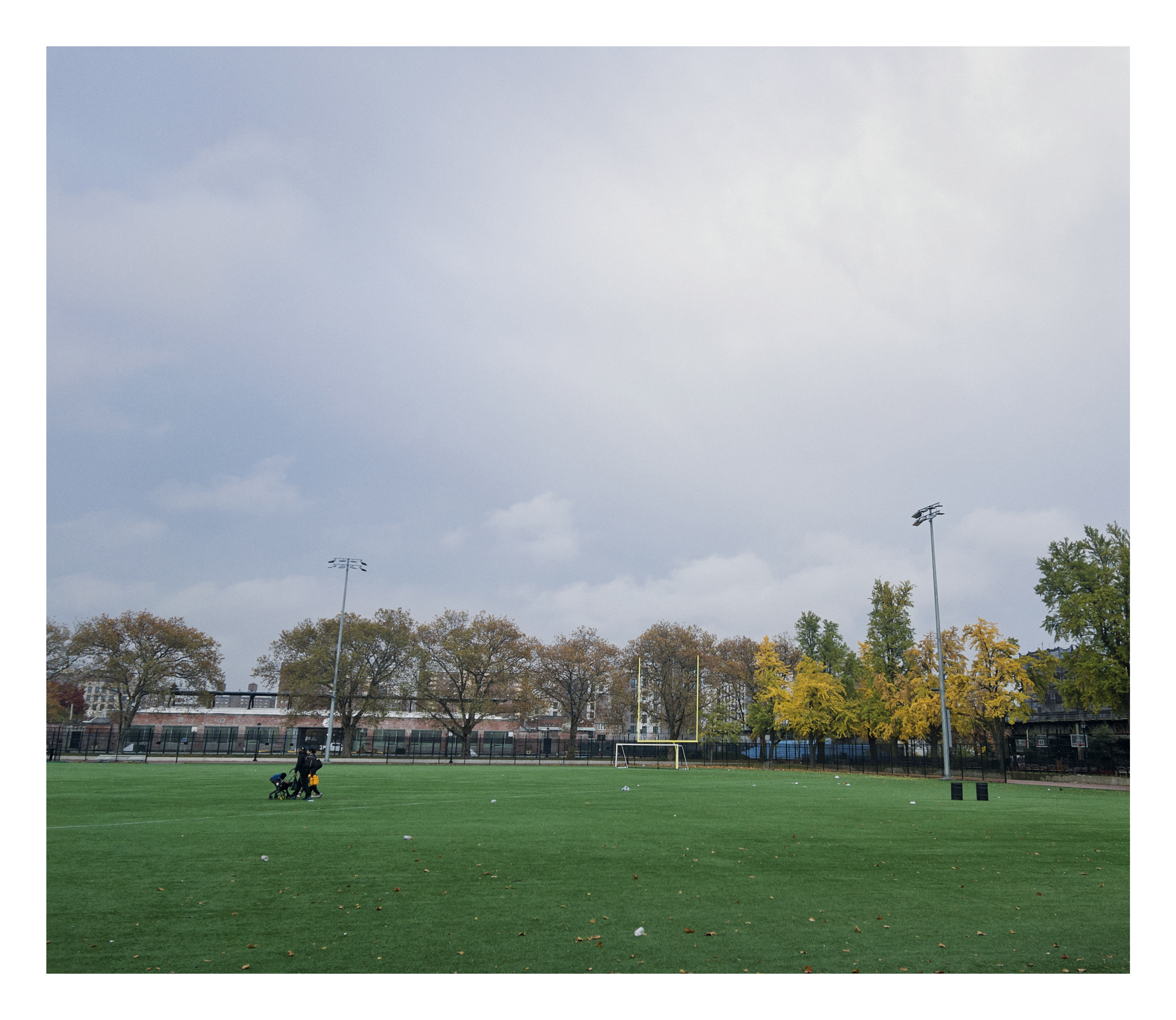 photograph of a couple walking a stroller across the bright green lawn of an athletic field; an overcast grey sky looms overhead, pierced by tall stadium lights