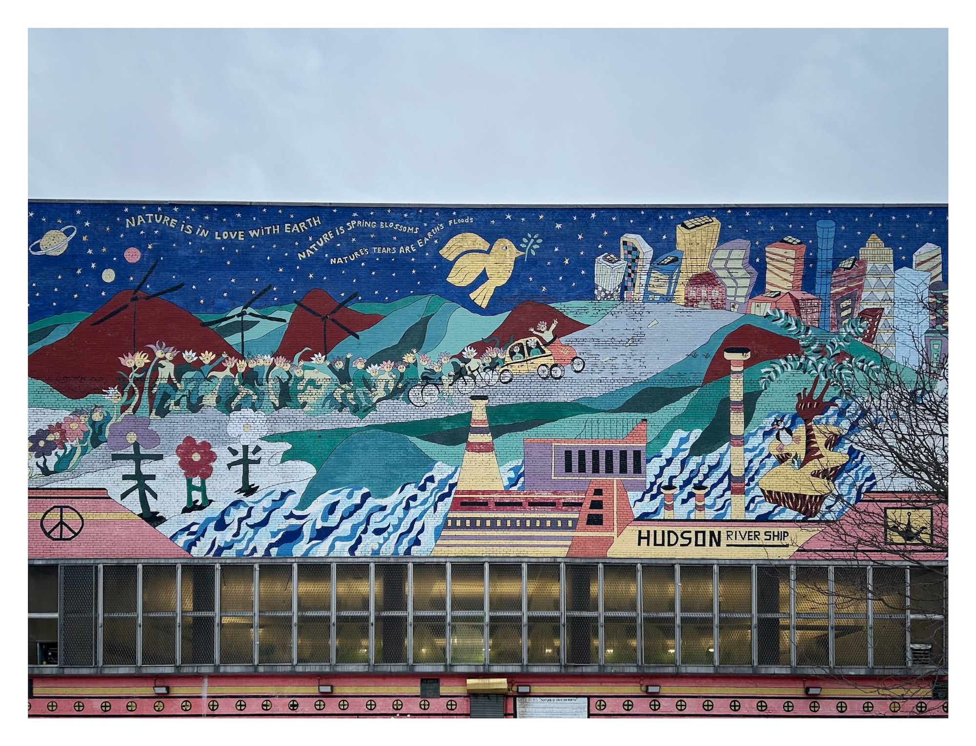 Mural depicting a people-, flower-, bike-, and car-filled road, flanked by brick red mountains and verdant rolling hills on one side, and Hokusai-inspired ocean waves on the other. A funky yellow factory is situated in the foreground, and a vibrant collection of city skyscrapers looms in the distance. "Nature is Love on Earth" by artist Duda Penteado and community