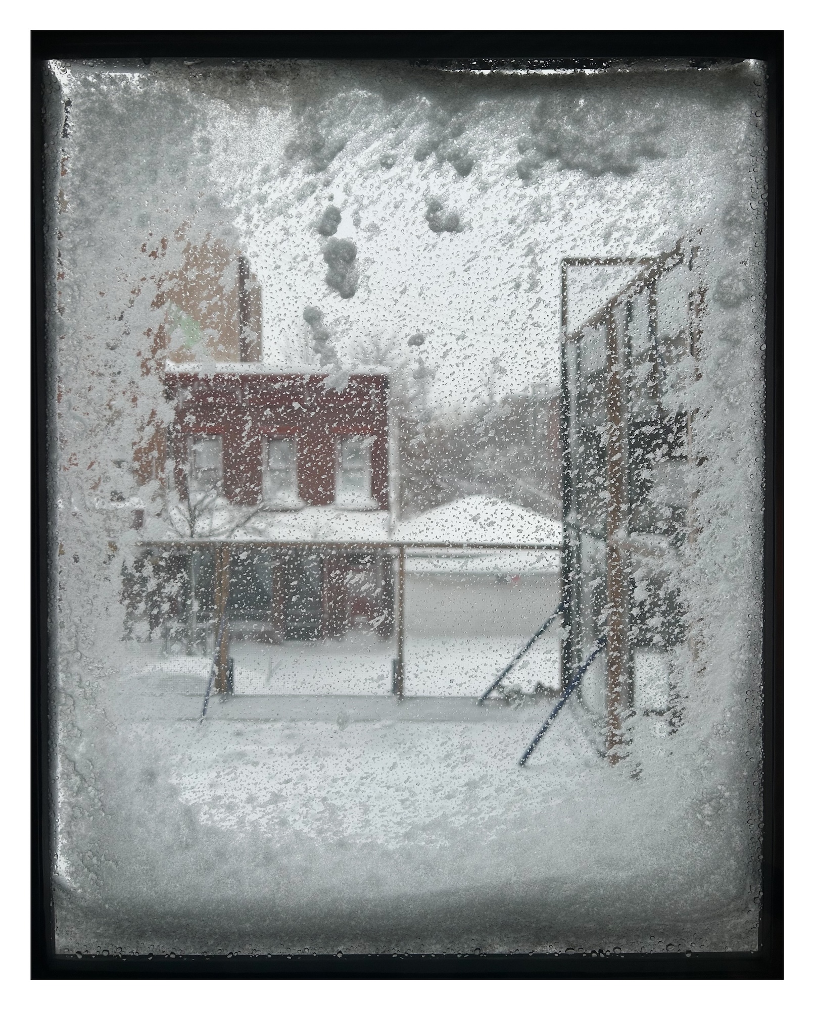 view outside toward a snow-covered street from inside a house through a window where wind-blown snow has stuck to the pane