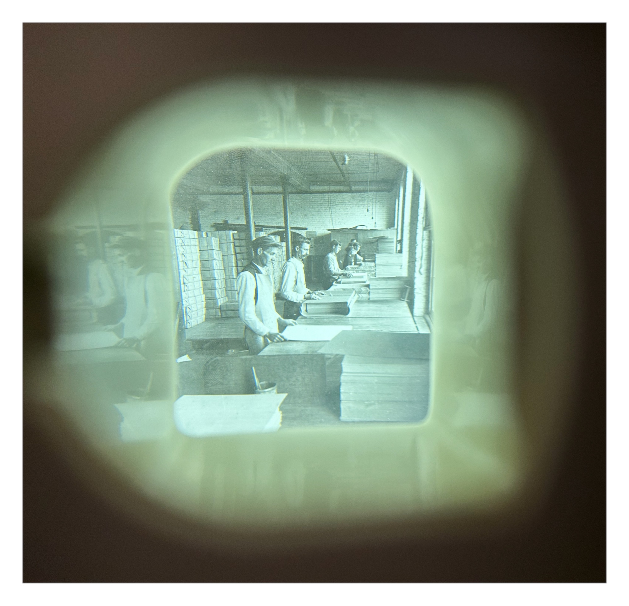 a view through one lens of a stereoscope to a photograph of men working in a papermaking shop