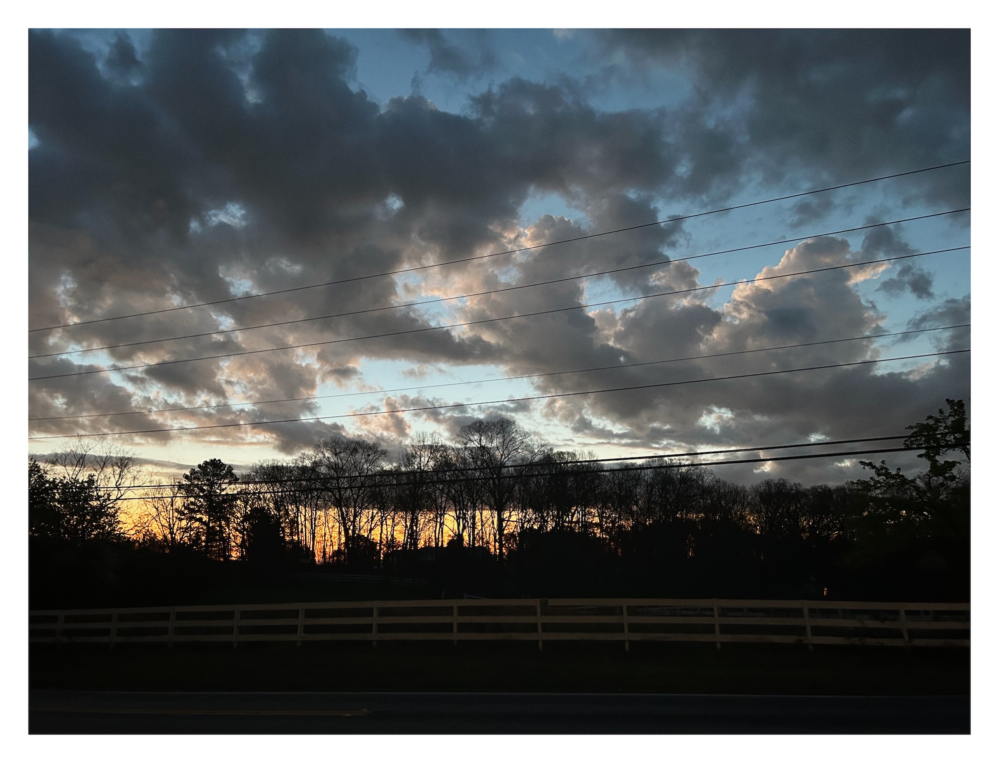 sunrise silhouettes a distant forest and lights the edges of clouds above a country road lined by a fence