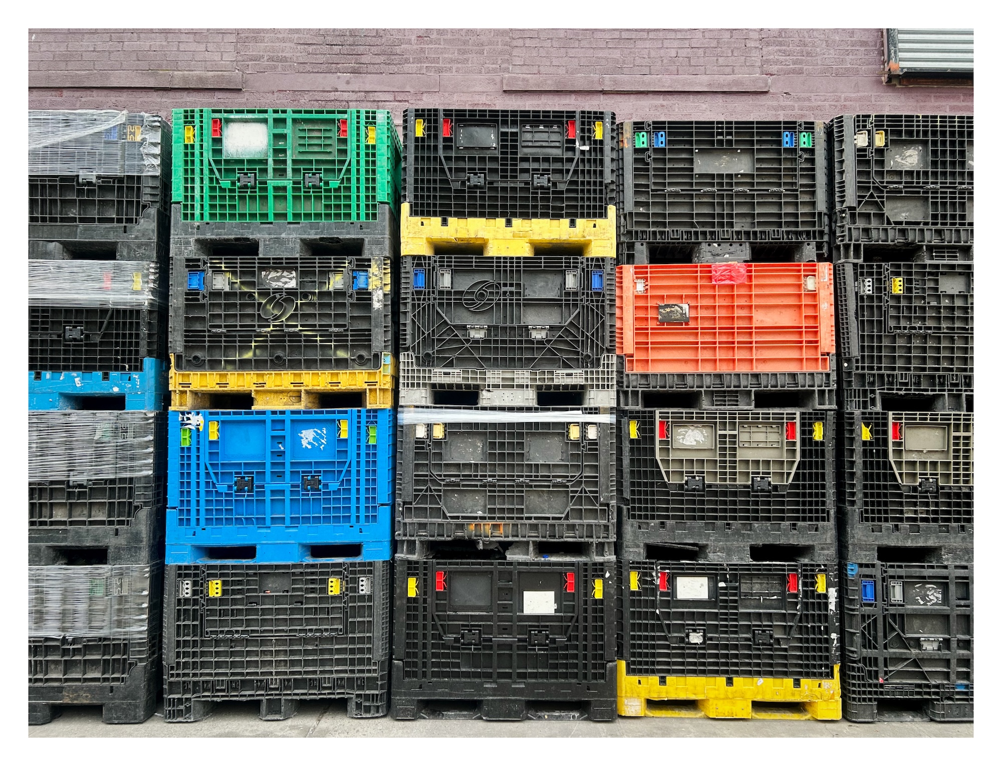large plastic shipping crates – mostly in black but with an occasional bright yellow, orange, blue, and green – are stacked in a grid outside a brick building