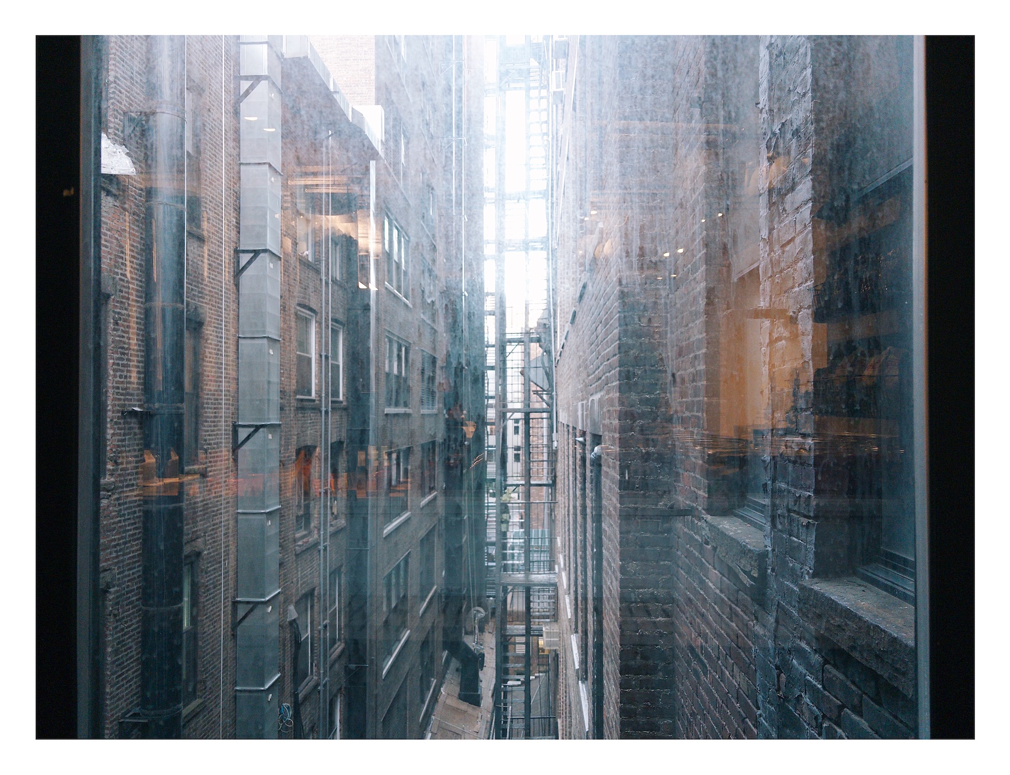 view of a narrow alley in Manhattan, lined with fire escapes mounted on brick walls, as seen through a dirty window