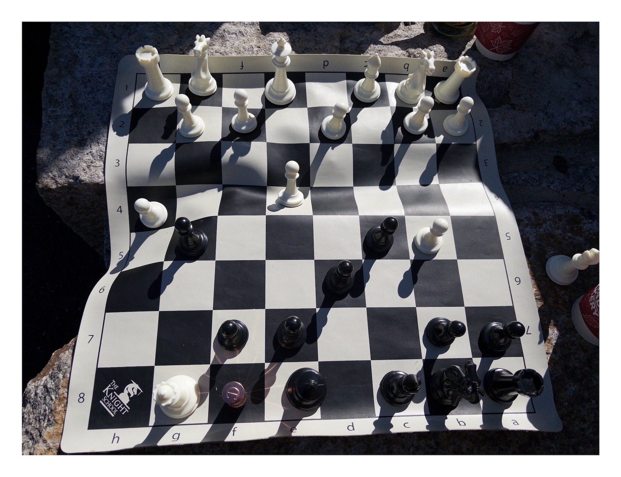 a plastic mat of a portable chess set ripples and flexes across an uneven surface, causing pieces on the black-and-white checkered surface to tilt in different directions in the morning sunlight. a tube of lipstick is being used as a bishop for black