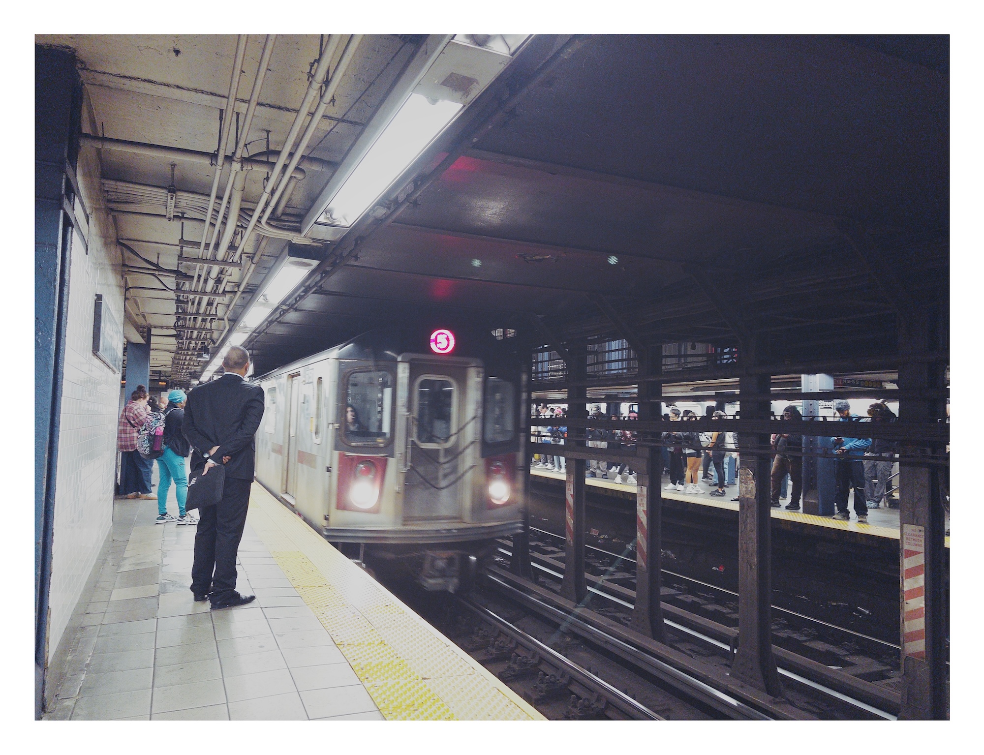 a man in a dark suit waits on a subway platform as the the NYC MTA 5 train pulls into the station